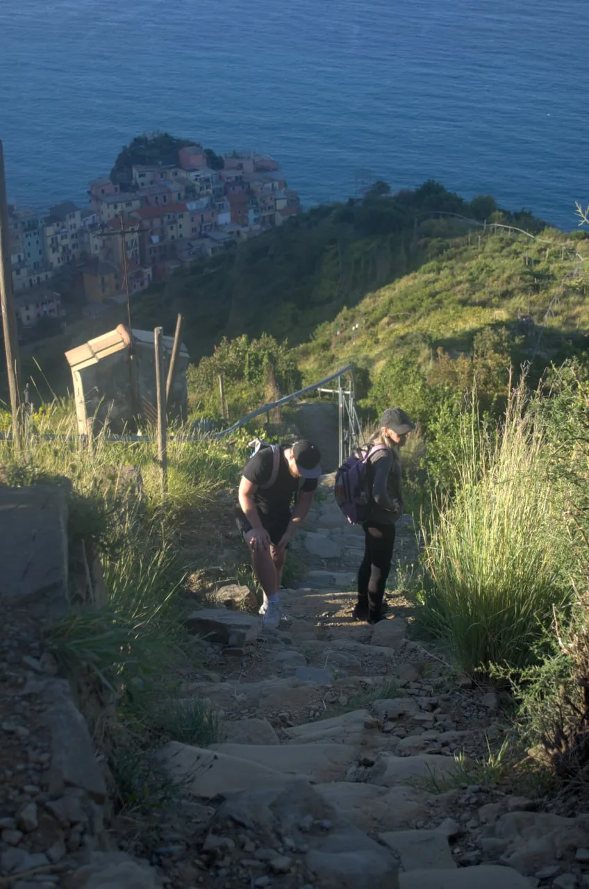 Hikers Rocky Climb J Photo Manarola To Corniglia Via Volastra. Two Hikers Pause On A Rocky Uphill Path, With A Coastal Village And Blue Sea Below.