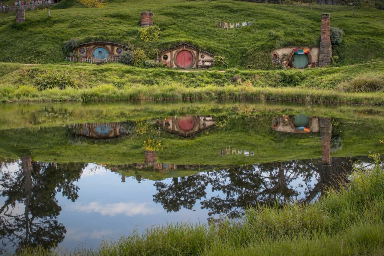 Blue, Red And Green Round Hobbit Doors Above A Pond, With Laundry Hanging Nearby And Reflections In The Water.