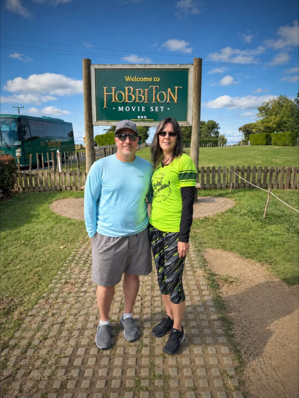 Two Visitors Beside The Welcome To Hobbiton Movie Set Sign, With A Tour Bus Behind Them.